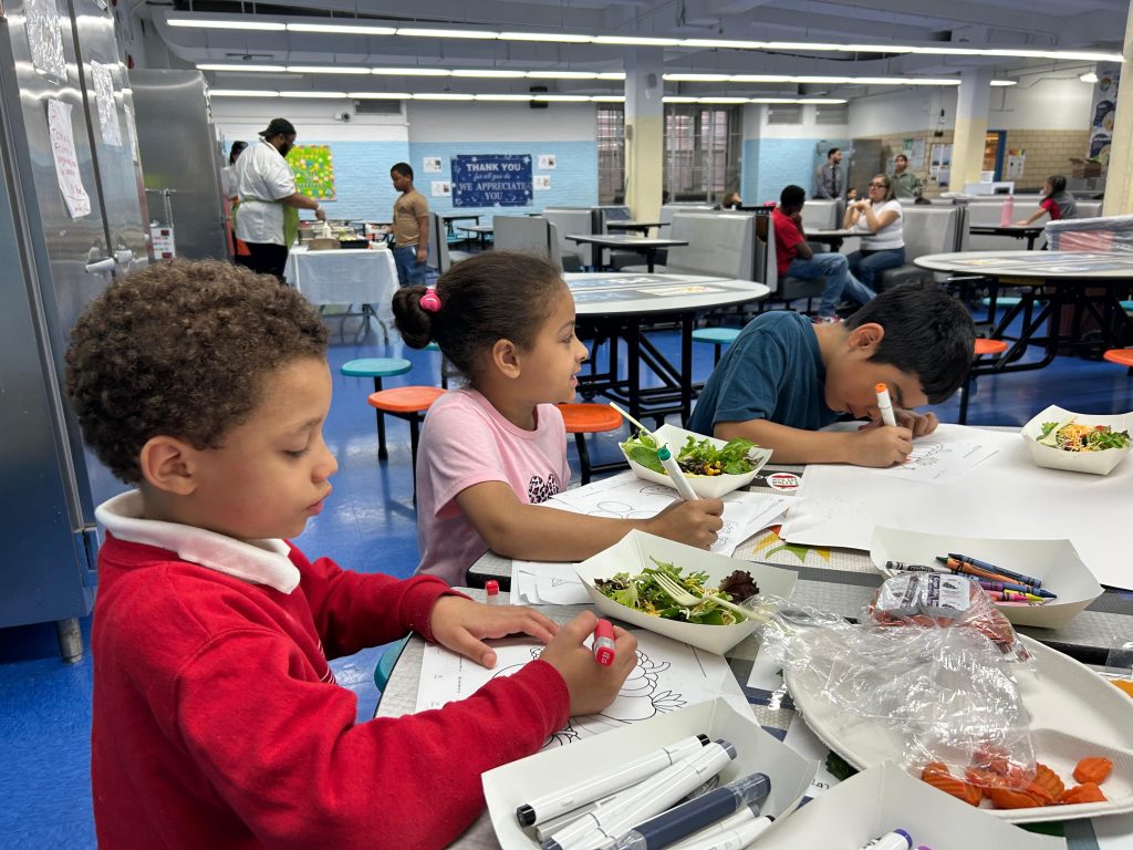 Three children sit at a table in the cafeteria. They have salads in front of them and are holding markers while coloring a worksheet.